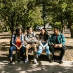 Four diverse college students sitting together outdoors on campus, smiling and talking between classes.