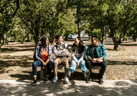 Four diverse college students sitting together outdoors on campus, smiling and talking between classes.