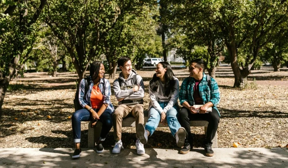 Four diverse college students sitting together outdoors on campus, smiling and talking between classes.
