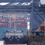 Container ships dock at a busy cargo port with stacked freight containers and towering loading cranes.