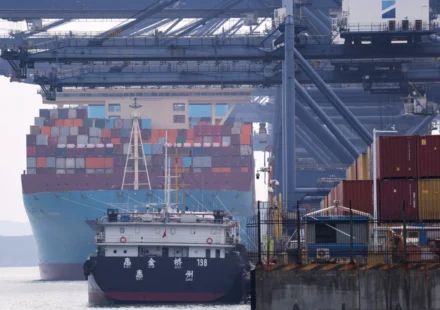 Container ships dock at a busy cargo port with stacked freight containers and towering loading cranes.