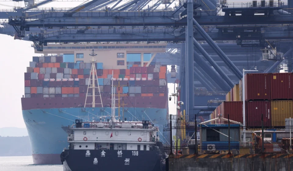 Container ships dock at a busy cargo port with stacked freight containers and towering loading cranes.