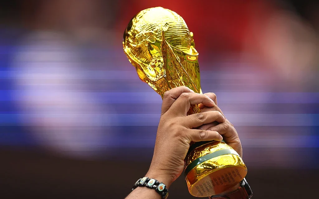 Hands raise the FIFA World Cup trophy before a blurred stadium crowd during an international football event.