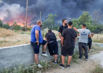 Residents watch thick smoke and advancing wildfire flames near a forest community in Greece at dusk.