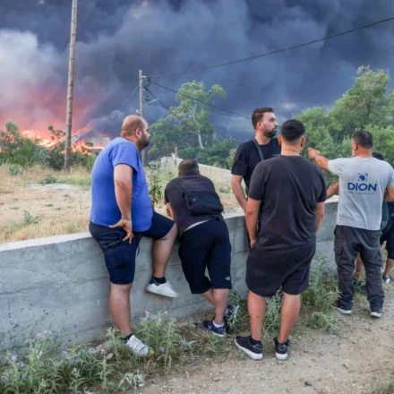 Residents watch thick smoke and advancing wildfire flames near a forest community in Greece at dusk.