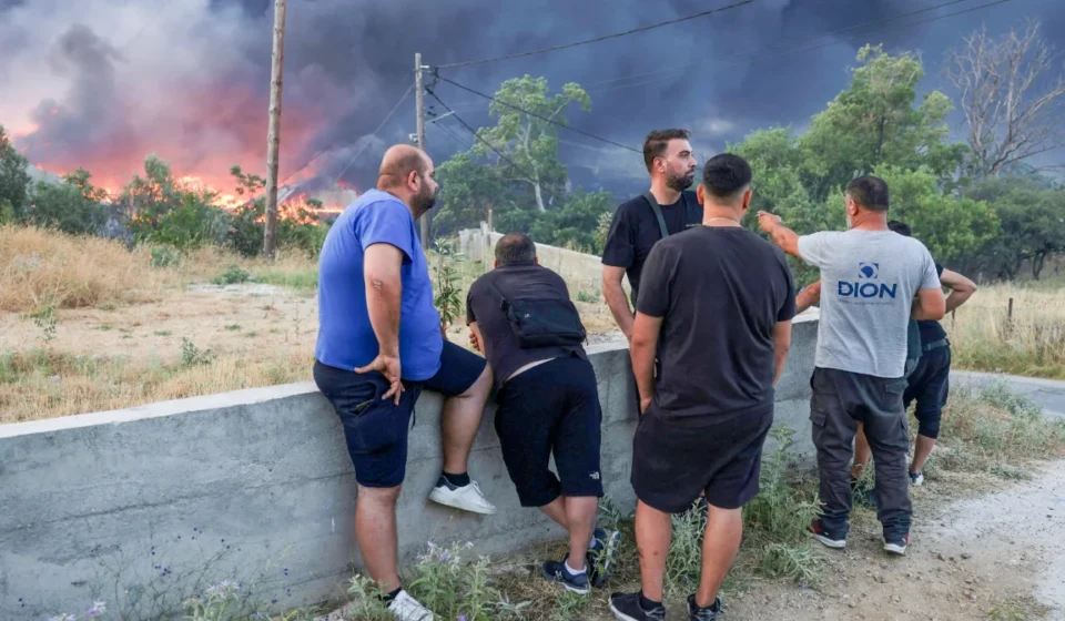 Residents watch thick smoke and advancing wildfire flames near a forest community in Greece at dusk.
