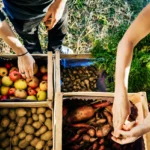 People selecting fresh local produce from wooden crates at a farmers market under warm morning sunlight.