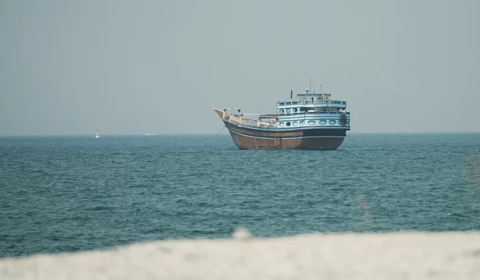 A traditional blue and white wooden dhow boat floating on calm blue ocean waters near a shoreline.