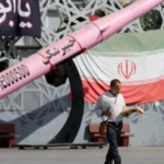 Man walks past Iranian flag and missile display amid rising regional military tensions and security alerts.