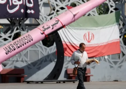 Man walks past Iranian flag and missile display amid rising regional military tensions and security alerts.