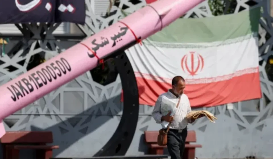 Man walks past Iranian flag and missile display amid rising regional military tensions and security alerts.