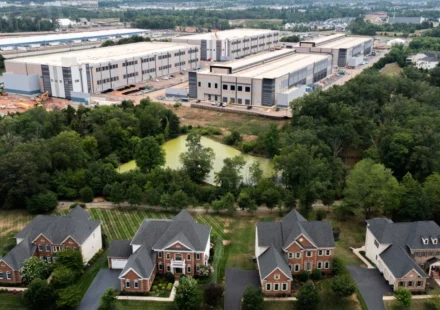 Aerial view of homes beside a large industrial data center complex surrounded by trees and greenery.