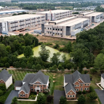 Aerial view of homes beside a large industrial data center complex surrounded by trees and greenery.