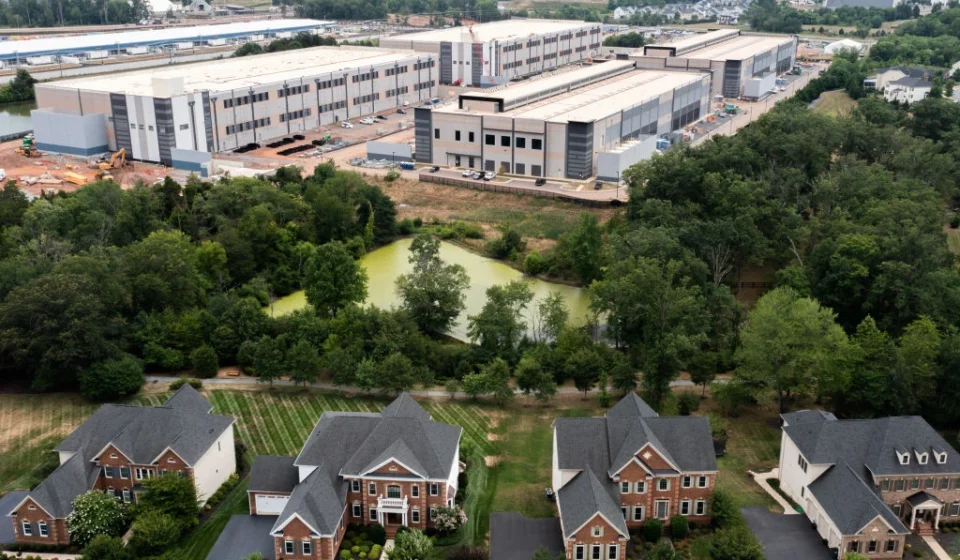 Aerial view of homes beside a large industrial data center complex surrounded by trees and greenery.
