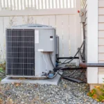 Outdoor residential HVAC condenser unit installed beside a house with utility connections and fence backdrop