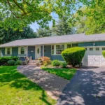 Blue single family home with attached garage, landscaped front yard, curved walkway, and mature trees.