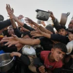 Crowd reaches for food as a volunteer distributes meals to families during a humanitarian aid distribution.