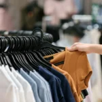 Clothing rack with neatly displayed shirts as a shopper selects a ribbed brown top in a retail store.