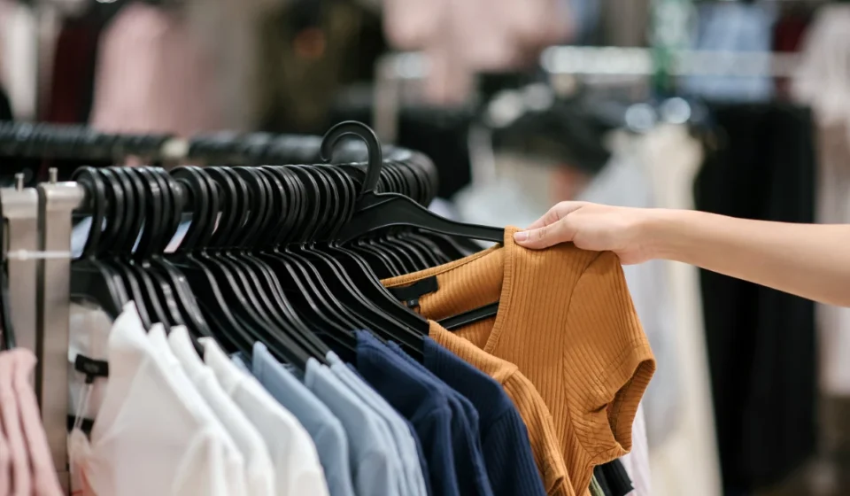 Clothing rack with neatly displayed shirts as a shopper selects a ribbed brown top in a retail store.
