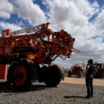 Large agricultural sprayer on display as a visitor examines modern farm equipment under a cloudy sky.