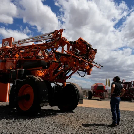 Large agricultural sprayer on display as a visitor examines modern farm equipment under a cloudy sky.