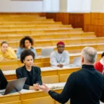 Professor lectures to diverse university students using laptops in a modern classroom during an AI education discussion.