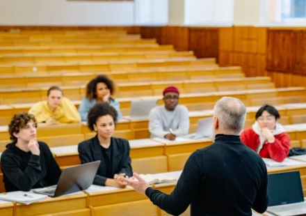 Professor lectures to diverse university students using laptops in a modern classroom during an AI education discussion.