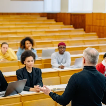Professor lectures to diverse university students using laptops in a modern classroom during an AI education discussion.