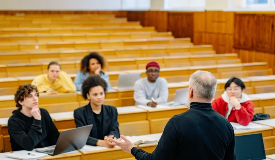 Professor lectures to diverse university students using laptops in a modern classroom during an AI education discussion.