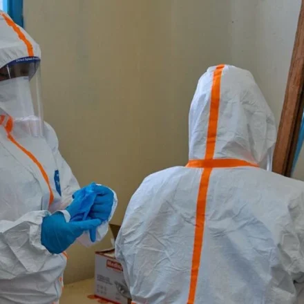 Healthcare workers wearing full protective suits prepare for Ebola response inside a medical facility room.