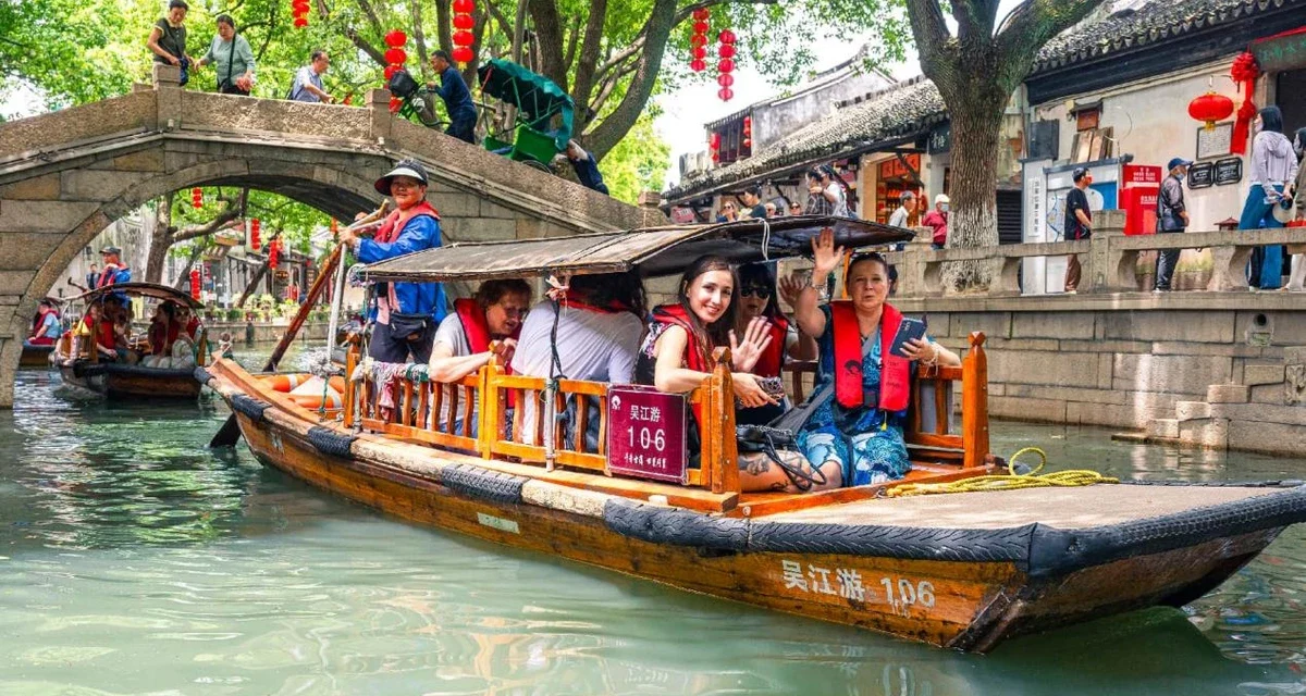 Traditional wooden boat carrying tourists through a scenic Chinese canal lined with historic buildings and trees.