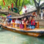 Traditional wooden boat carrying tourists through a scenic Chinese canal lined with historic buildings and trees.
