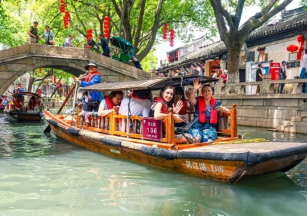 Traditional wooden boat carrying tourists through a scenic Chinese canal lined with historic buildings and trees.