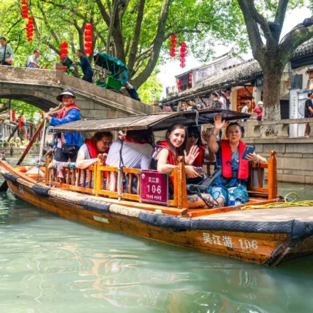 Traditional wooden boat carrying tourists through a scenic Chinese canal lined with historic buildings and trees.