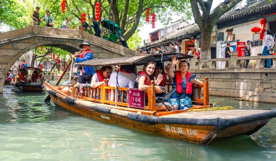 Traditional wooden boat carrying tourists through a scenic Chinese canal lined with historic buildings and trees.
