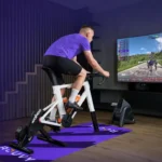 Man riding a smart indoor bike while Zwift displays a virtual cycling route on a large screen.
