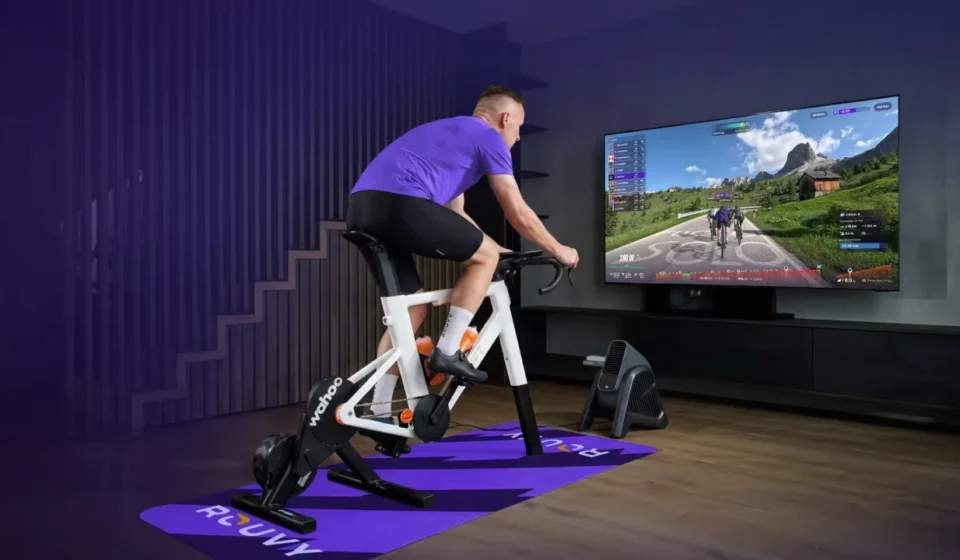 Man riding a smart indoor bike while Zwift displays a virtual cycling route on a large screen.