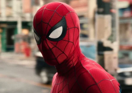 Spider Man in his iconic red suit stands on a city street, looking over his shoulder in daylight.