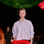 Model walks runway in a striped Chanel shirt during a fashion show showcasing the brand’s chic style.