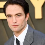 Robert Pattinson poses in a gray suit and tie at a red carpet event, gazing toward the camera ahead.