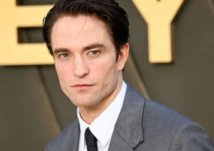 Robert Pattinson poses in a gray suit and tie at a red carpet event, gazing toward the camera ahead.
