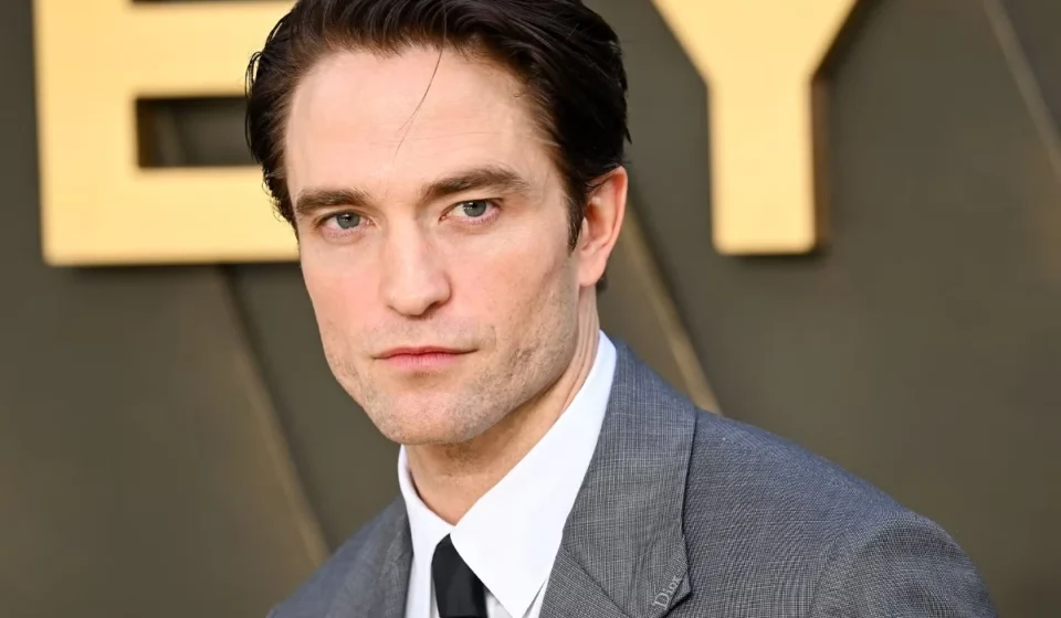 Critical Acclaim Surges for Robert Pattinson’s Portrayal in A24’s Primetime Robert Pattinson poses in a gray suit and tie at a red carpet event, gazing toward the camera ahead.