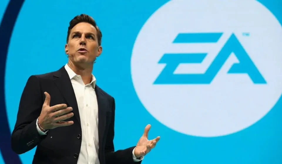 Electronic Arts Finalizes $55 Billion Buyout and Delists EA executive speaks on stage with the Electronic Arts logo displayed on a large screen behind him.