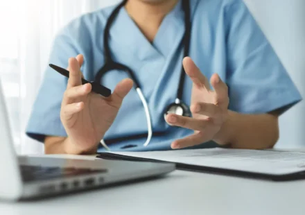Doctor conducts a telehealth consultation while discussing patient care using a laptop and clipboard.