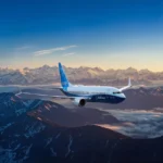 Boeing passenger jet flying above snow covered mountain peaks during sunrise with clear skies and clouds below