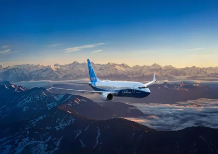 Boeing passenger jet flying above snow covered mountain peaks during sunrise with clear skies and clouds below