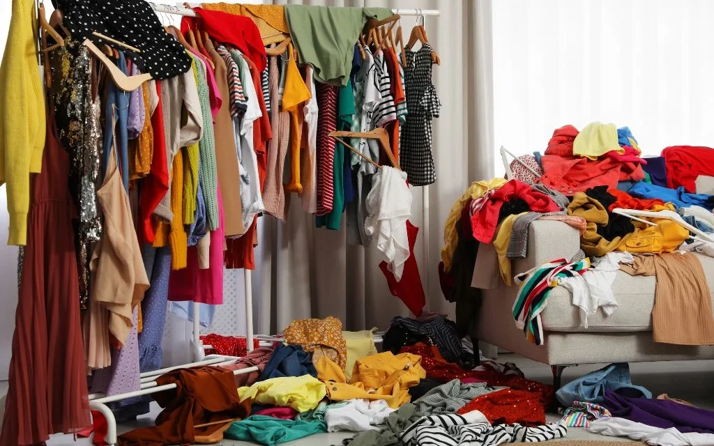 Clothing fills racks and piles in a crowded room, highlighting fashion waste and excess production.