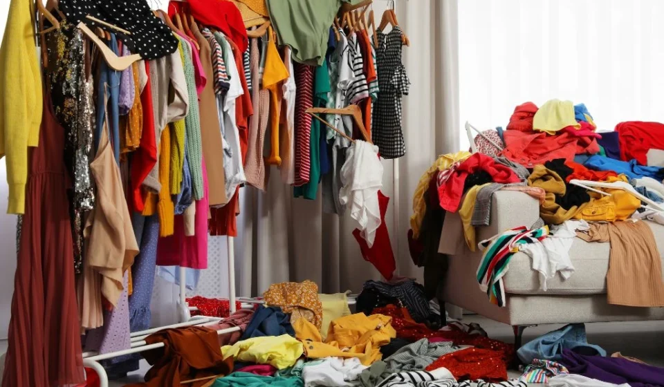 Global Fashion Accord Banishes Synthetic Microfiber Waste Clothing fills racks and piles in a crowded room, highlighting fashion waste and excess production.