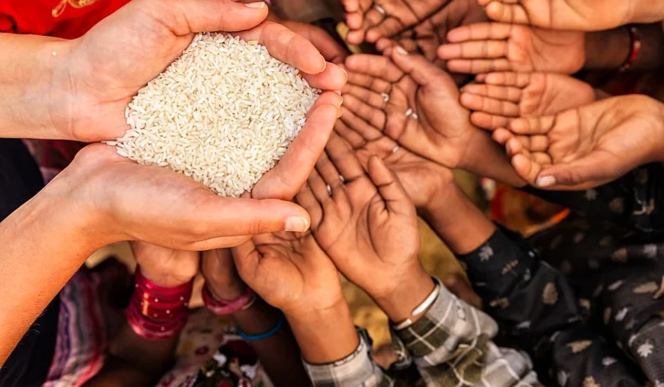 People reach out with empty hands toward a bowl of rice, highlighting hunger and food insecurity.