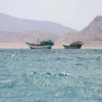Boats on blue waters near rocky mountains in the Strait of Hormuz beneath hazy skies at daybreak sea